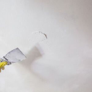 worker's hand in yellow gloves holding putty knife patching a hole with spatula with plaster or putty in white wall. Renovation and repair process, remodeling interior of room at apartment building